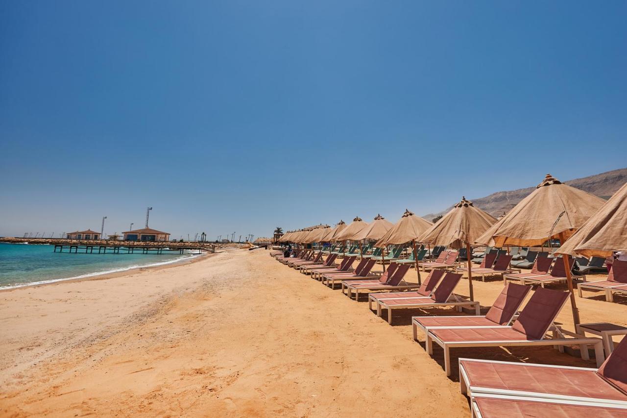 Tolip Resort El Galala Majestic Ain Sukhna Exterior foto The photo shows a scenic beach with a stretch of golden sand under a clear blue sky. There are several beach loungers arranged in rows, each accompanied by thatched umbrellas providing shade. In the background, there is a wooden pier extending into t