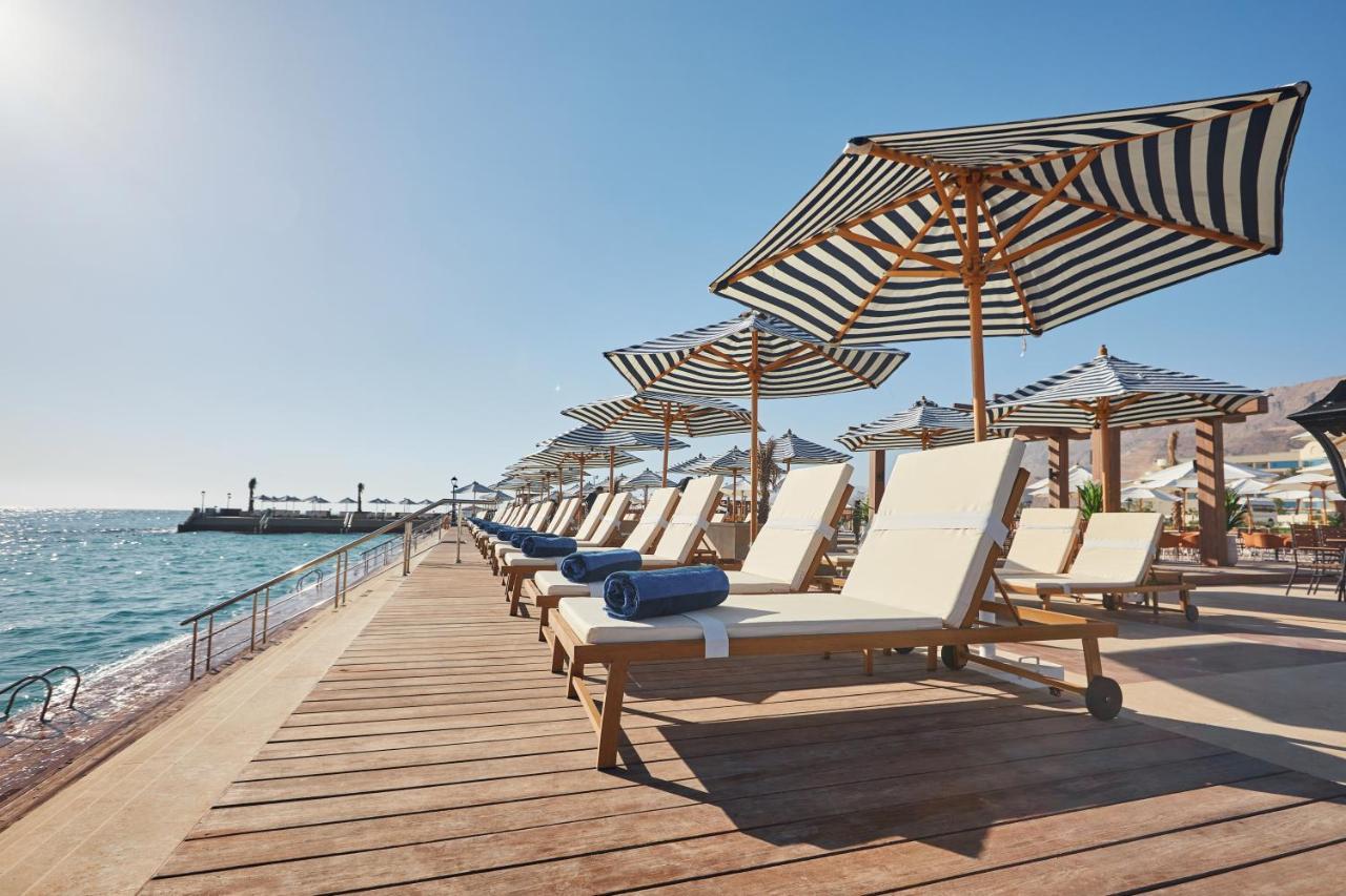 Tolip Resort El Galala Majestic Ain Sukhna Exterior foto The photo depicts a serene beachside setting featuring a row of sun loungers arranged on a wooden deck. Each lounger is equipped with a folded towel and shaded by striped umbrellas. In the background, the water sparkles under bright sunlight, and the