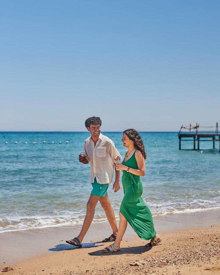 Tolip Resort El Galala Majestic Ain Sukhna Exterior foto The photo shows a couple walking along a beach. The man is wearing a white short-sleeved shirt and green shorts, while the woman is dressed in a flowing green dress. They both appear to be enjoying the pleasant weather, with the ocean in the backgrou