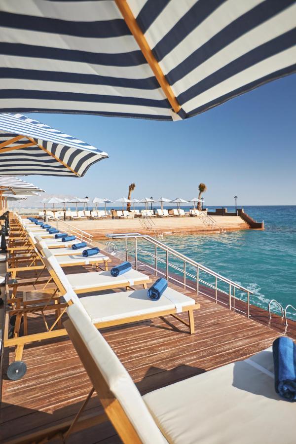 Tolip Resort El Galala Majestic Ain Sukhna Exterior foto The photo depicts a serene outdoor area by the waterfront. There are several lounge chairs arranged in a row, each equipped with blue towels. Large, striped umbrellas provide shade over some of the chairs. In the background, there is a wooden deck th